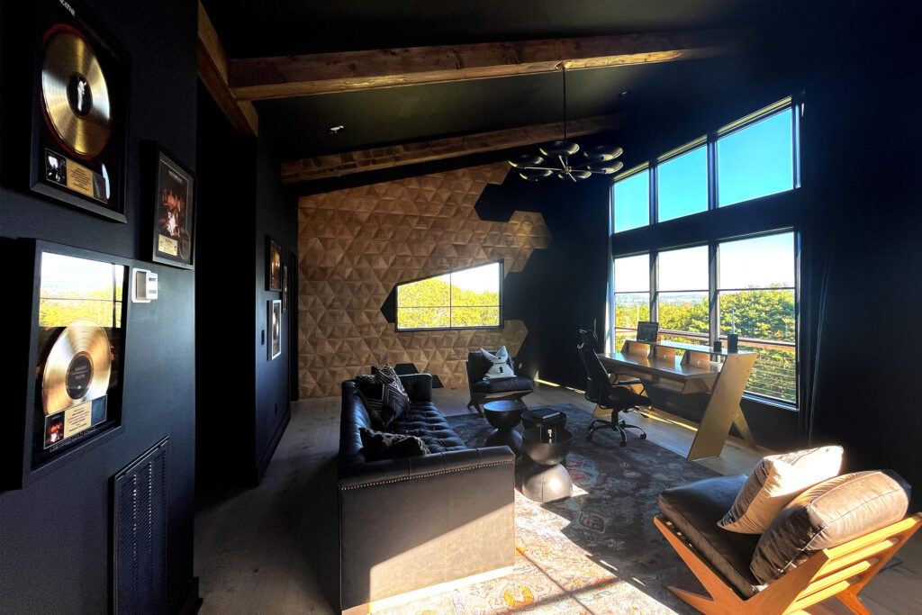 A wide interior view of a luxury dark-walled home studio featuring a large window wall, a modern desk setup, and a geometric oak wooden panel accent wall.