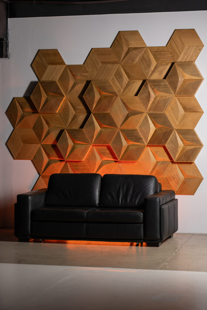 A wide interior shot showcasing a black leather sofa and a custom wall installation made of hexagonal 3D acoustic wooden panels with orange backlighting.