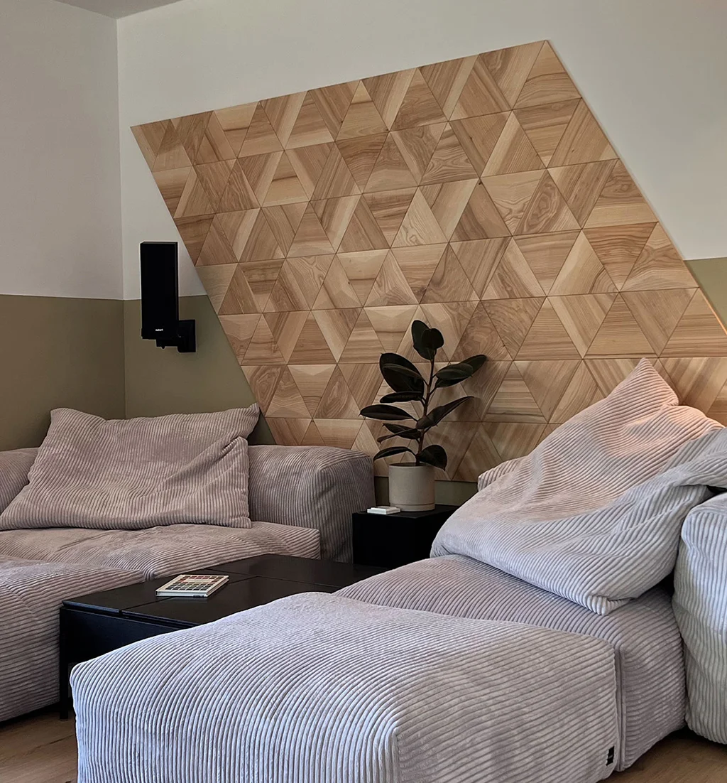 Minimalistische Wanddekoration aus flachen Eichenholz-Dreiecken in einem Wohnzimmer mit einem hellgrauen Cordsofa und einer Zimmerpflanze.