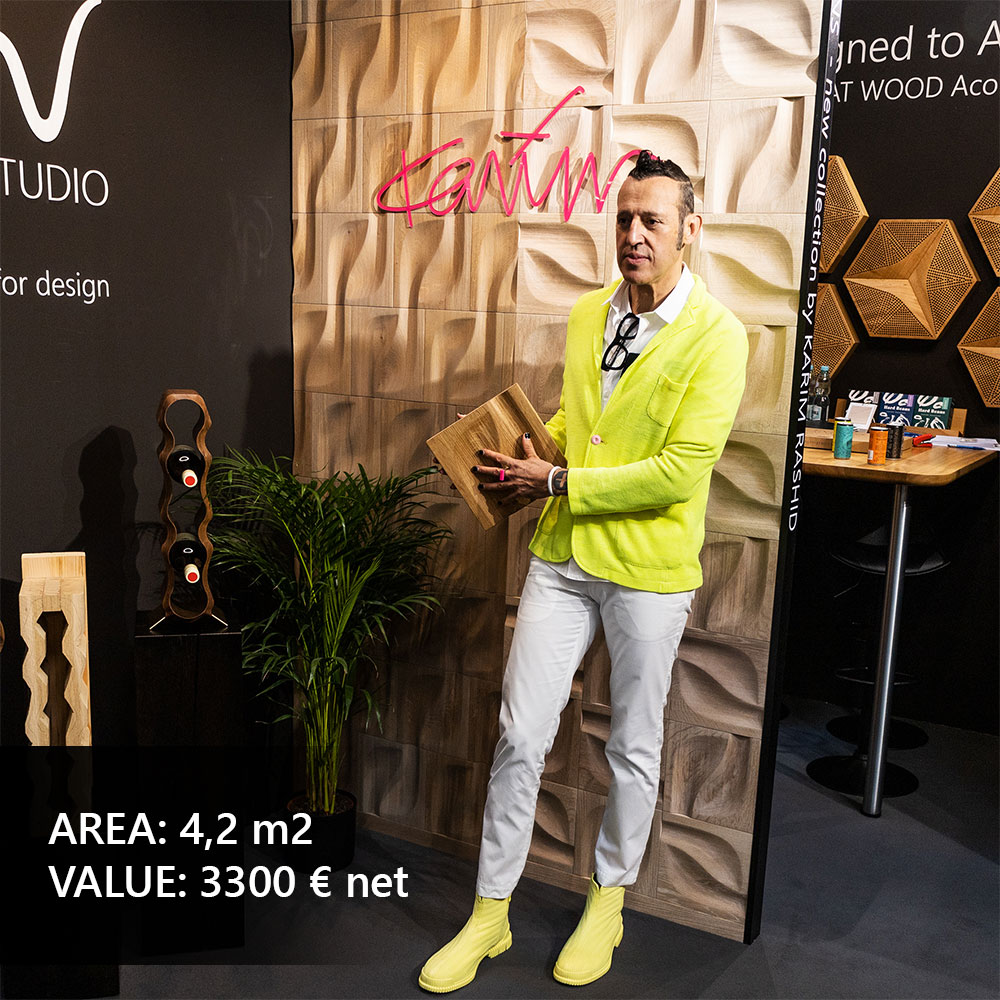 Karim Rashid präsentiert ein 3D-Eichenpaneel vor einer Ausstellungswand mit organischen Formen auf einer Designmesse.