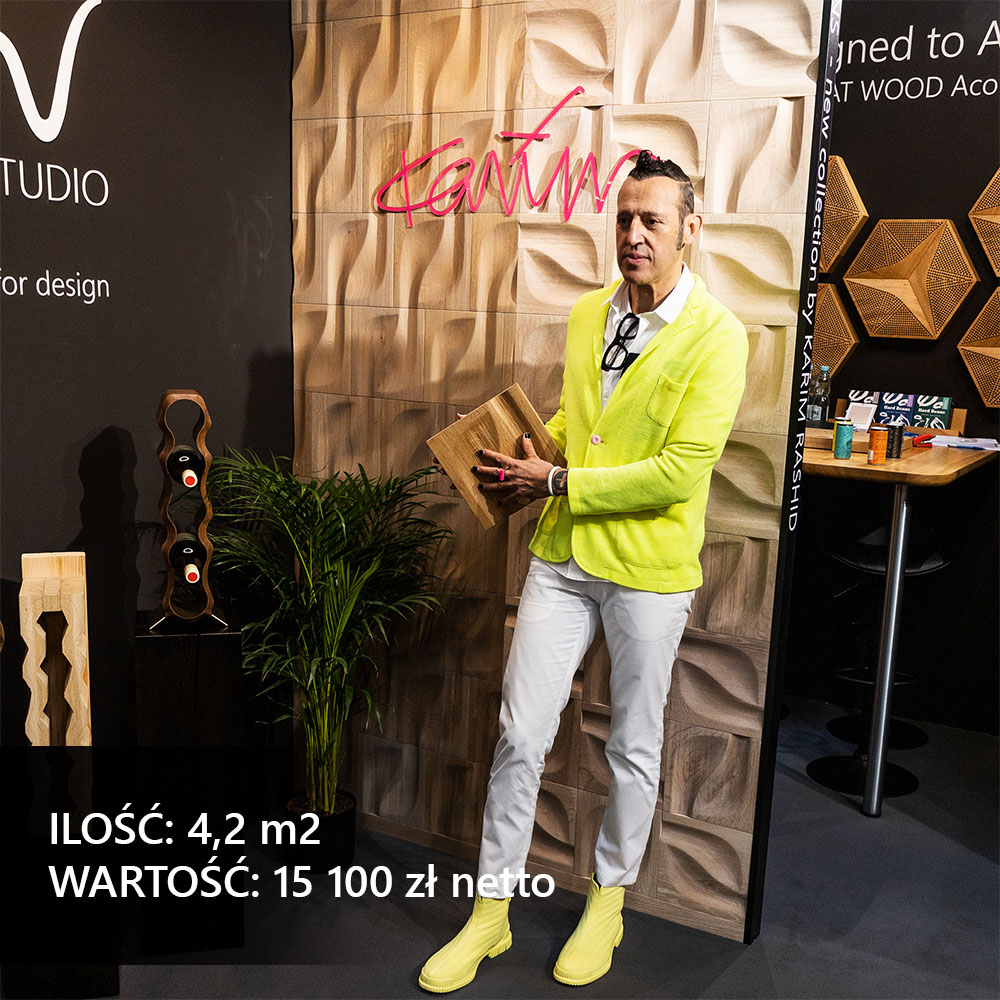 Karim Rashid prezentujący dębowy panel 3D na tle ściany ekspozycyjnej o organicznych kształtach podczas targów designu.