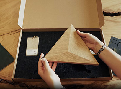 Dłonie wyjmujące dębowy panel trójwymiarowy z serii Pyramid z otwartego pudełka z próbkami FORM AT WOOD w domowym wnętrzu.